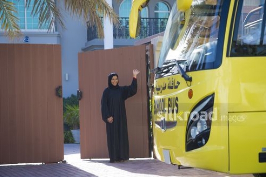 Middle Eastern mother waving outside of her house in front of a school bus|-