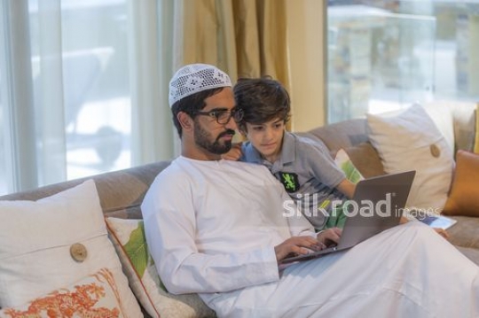 Arab man sitting with the young kid on the couch using the laptop|