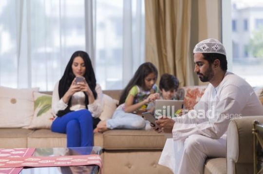 Middle Eastern man on the device with his family in the background on their own devices|