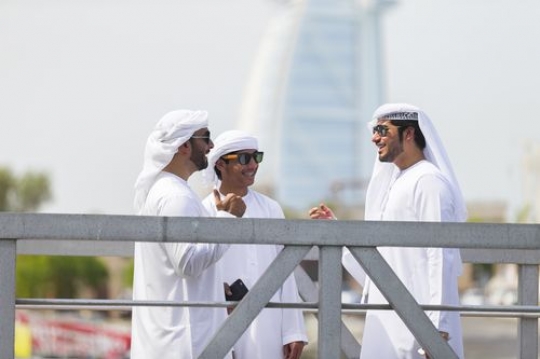 Three arab men standing by the dock dressed in the traditional UAE attire talking to each other|