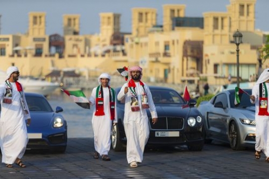Middle Eastern men walking and three luxury cars behind them during the UAE national Day Parade|-