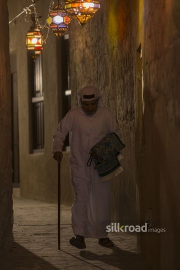 Arab man carrying a prayer rug while walking through the alleyway of Ramadan Lanterns|-