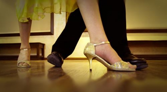 dancing womans feet in gold sandals on brown floor with man foot moving away